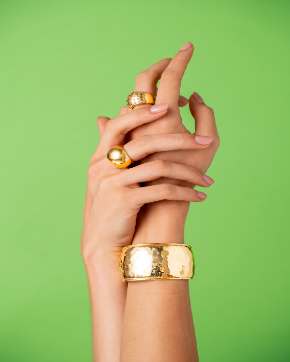 Mano femminile con gioielli in oro (anelli e bracciale) su sfondo verde, simbolo di lusso sostenibile e gioielleria etica
